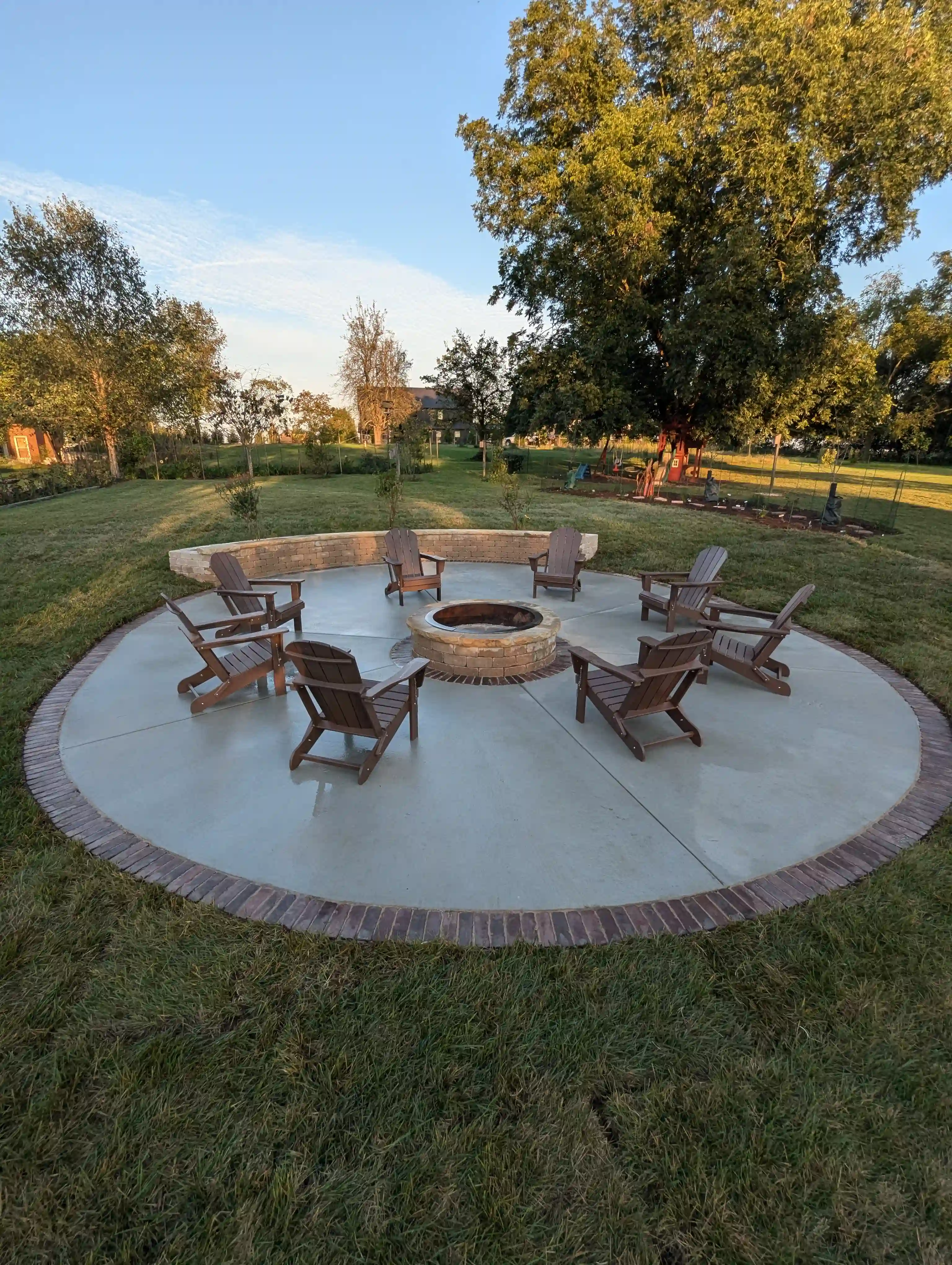 circular firepit hangout area with with multiple chairs. the concrete has red brick lining the perimeter and a stone retaining wall