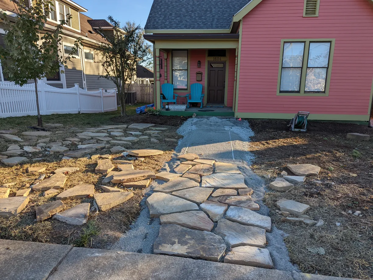 a progress picture redoing a walkway from the sidewalk to the frontdoor of a home. there are a lot of flagstone scattered in the frame ready to be placed together
