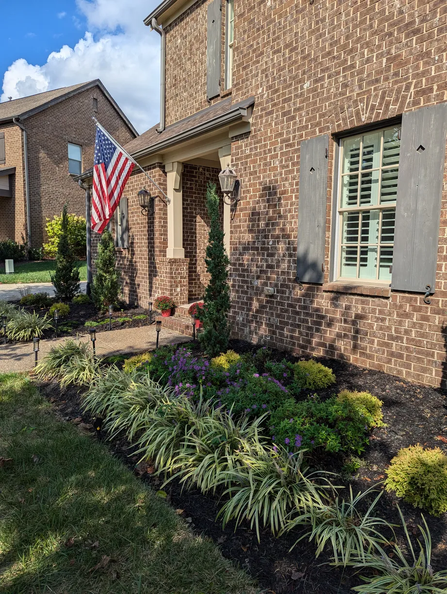 american flag perched on a home with a beautiful foundation bed filled with many small plants and walkway lights